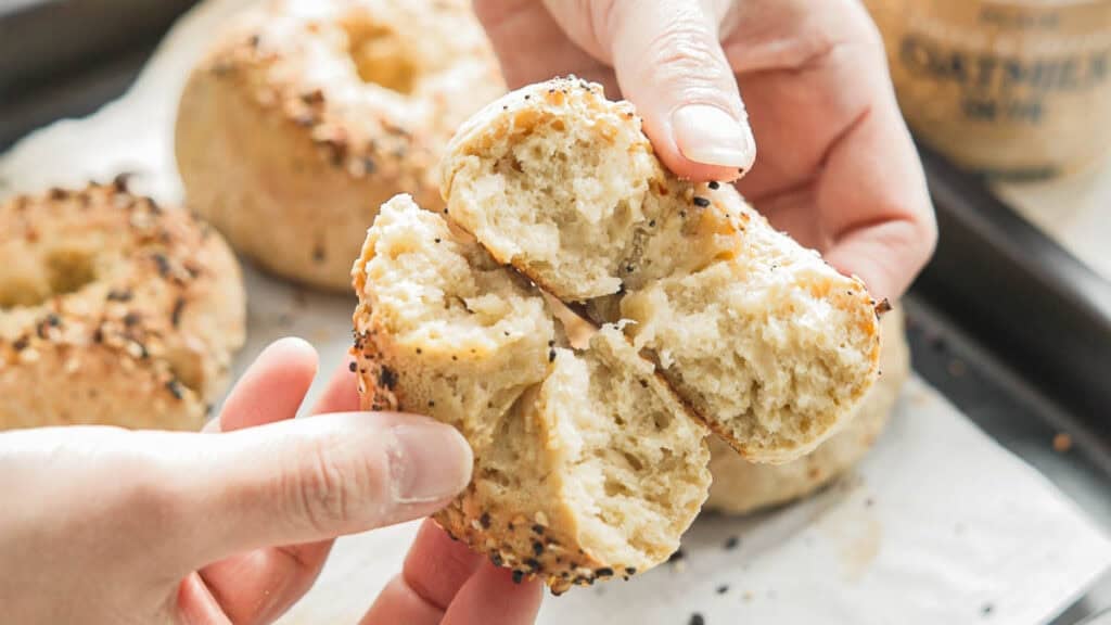 Hands tearing apart a freshly baked, sesame seed-crusted 3-ingredient bagels, revealing a soft, fluffy interior. Other bagels on a parchment-lined tray are visible in the background.